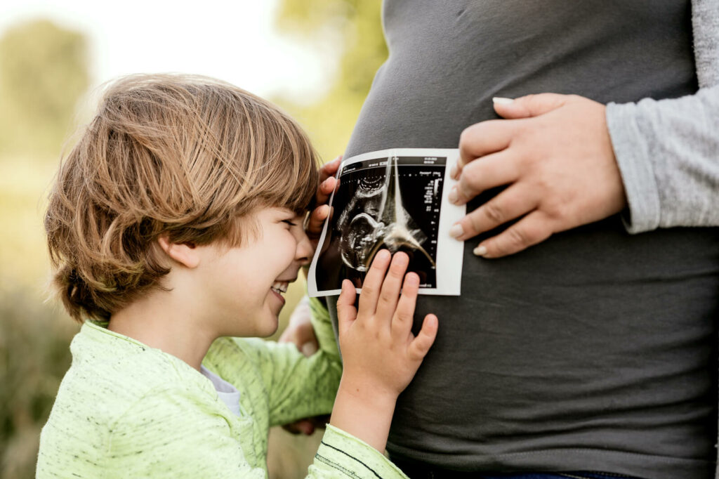 Ein Kind berührt mit geschlossenen Augen lächelnd ein Ultraschallbild, das von einer erwachsenen Hand an einen Babybauch gehalten wird.