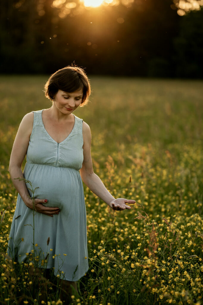 Eine Schwangere steht im Sonnenuntergang auf einer Blumenwiese. Sie hält mit einer Hand lächelnd ihren Babybauch.