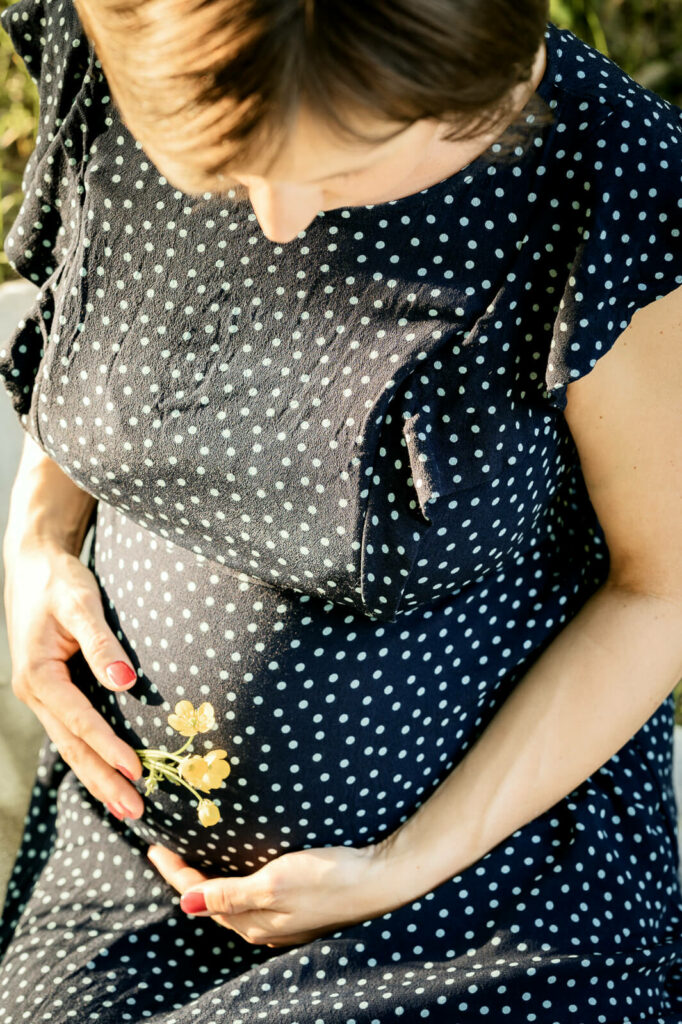 Eine Schwangere hält eine gelbe Blume vor ihren Babybauch. Sie trägt ein gepunktetes Kleid.