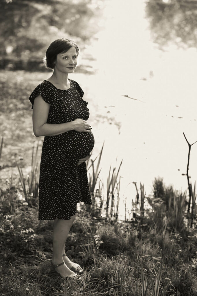 Eine Schwangere steht vor einem See. Sie hält mit beiden Händen ihren Babybauch. Schwarzweiß-Aufnahme