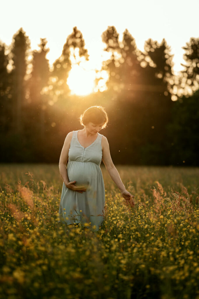 Eine Schwangere steht im Sonnenuntergang auf einer Blumenwiese. Sie hält mit einer Hand lächelnd ihren Babybauch und berührt mit der anderen das Gras