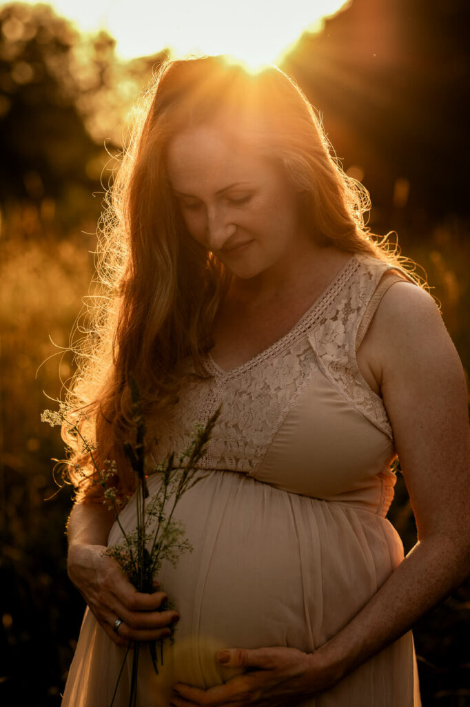 Eine Schwangere steht im Sonnenuntergang. Sie hält einen Strauß Gräser an ihren Babybauch.