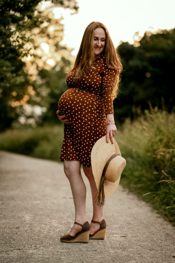 Eine Schwangere im gepunkteten Kleid auf einem Weg im Grünen. Sie dreht asich gerade zur Kamera und hält einen Strohhut in den Händen