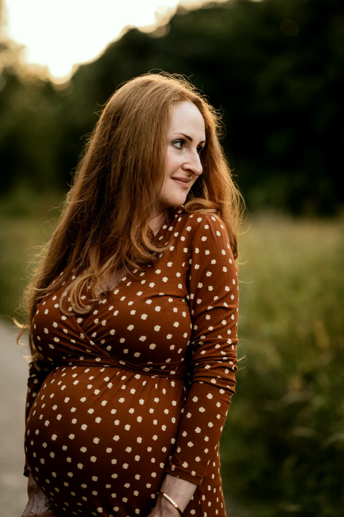 Portrait einer Schwangeren mit langen roten Haaren im braun-weiß gepunkteten Kleid. Sie blickt über ihre Schulter und a erkennt ihren Babybauch