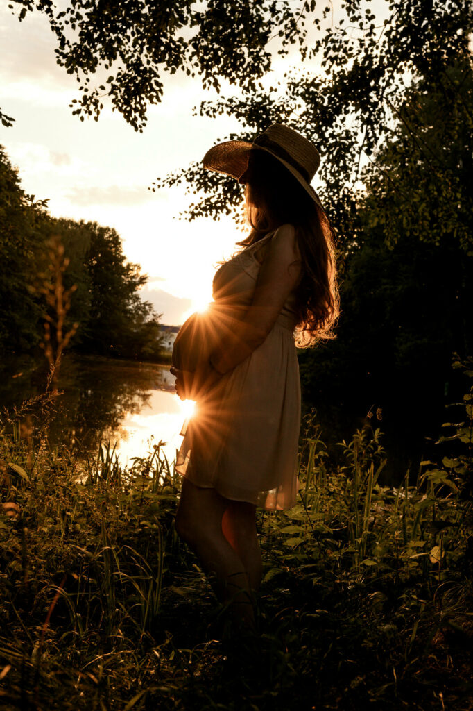 Eine Schwangere steht im Sonnenuntergang. Sie lehnt an einem Baum udn hat die Hände auf ihren Babybauch gelegt. Oberhalb und unterhalb des Bauches strahlen Sonnensterne