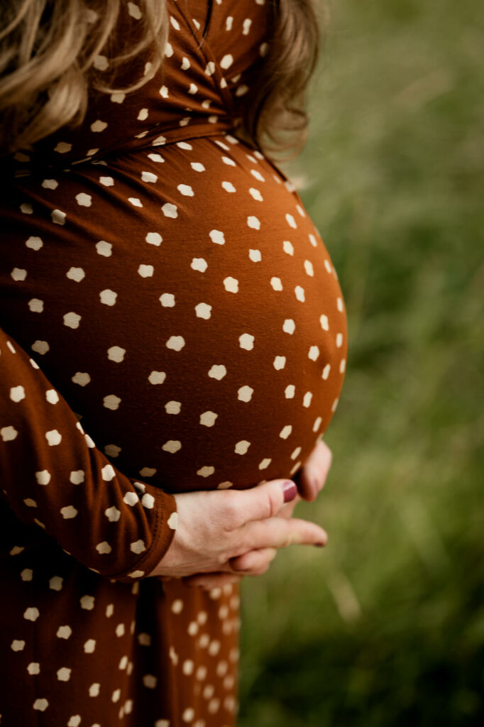 Ein Babybabybauch in einem braun-weiß gepunkteten Kleid. Die Schwangere umfasst ihn von unten mit den Händen