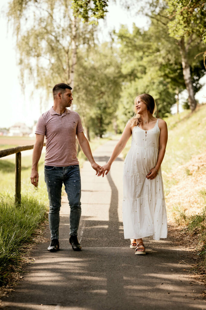 Ein Paar geht Hand in Hand einen Weg entlang. Die schwangere Frau im weißen Kleid hält mit der anderen Hand ihren Babybauch